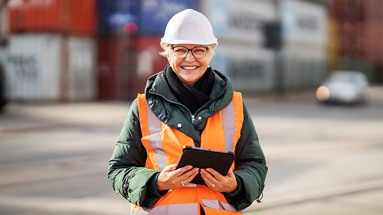 Portrait of successful female engineer with digital tablet at container warehouse. Senior woman in protective workwear looking at camera and smiling.