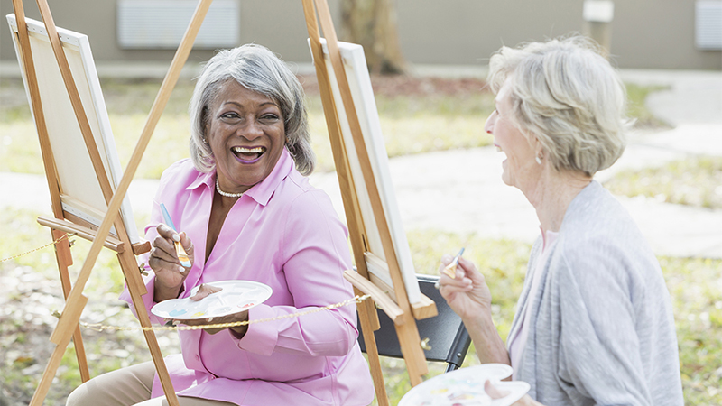 Two women smiling and laughing as they paint outdoors