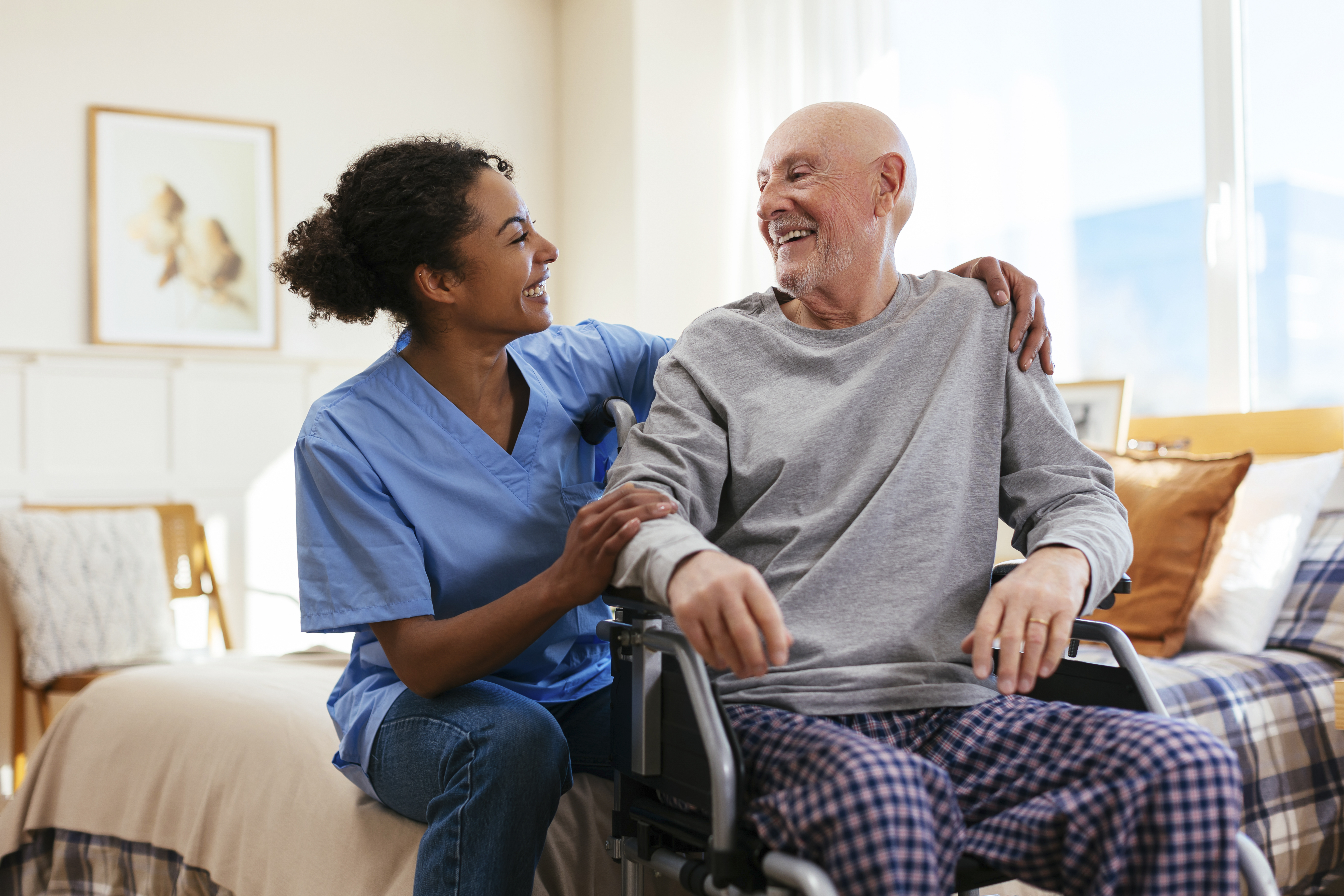 Happy nurse with senior man sitting on wheelchair in bedroom at home