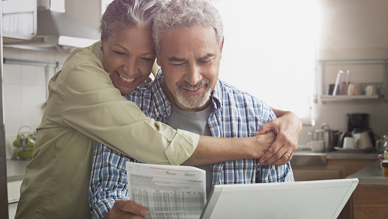 Older Hispanic woman holds her older Hispanic husband while they look over a medical bill together in a kitchen.