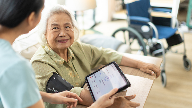 Woman Asian nurse taking blood pressure of a senior woman patient at home. Home care healthcare professional using digital tablet.