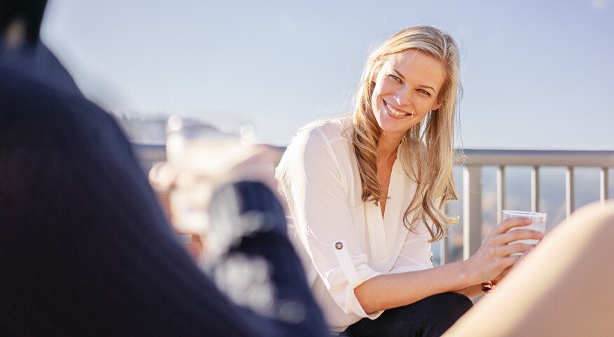 Woman with blonde hair, holding a drink and looking at man on balcony