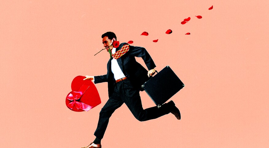 Man in suit running with rose between his teeth and heart shaped box of chocolates in his hand.