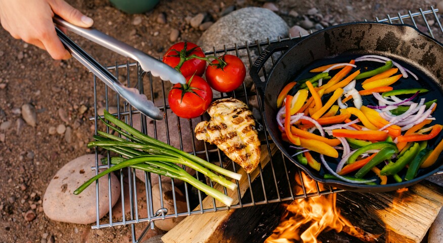 Hand holding tongs while cooking vegetables and chicken over an open fire
