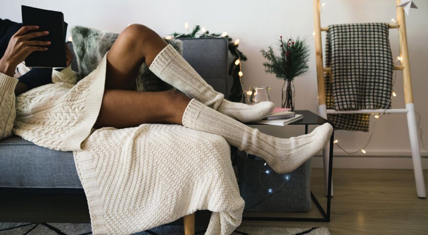 Woman lounging on a couch with Christmas lights in comfortable clothing reading a tablet