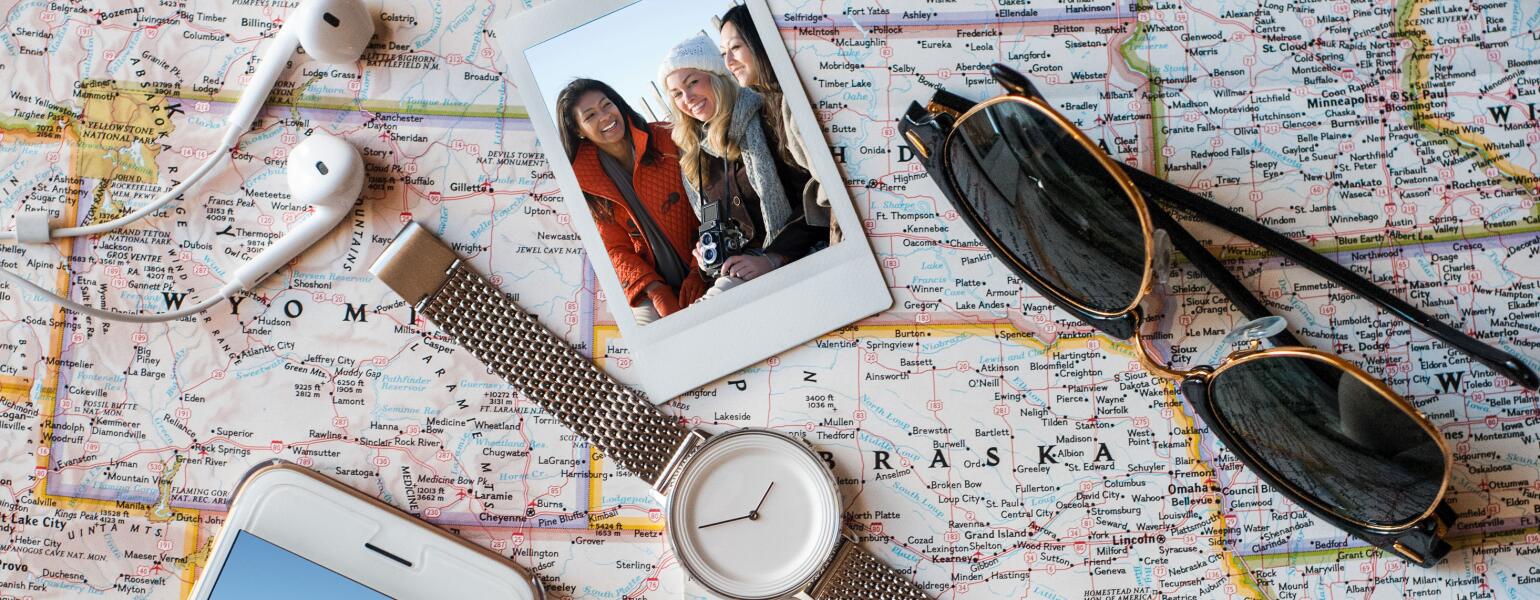 Photo of a U.S. map with miscellaneous travel items on top like a watch, sunglasses, headphones, money, cell phone and polaroid photo.