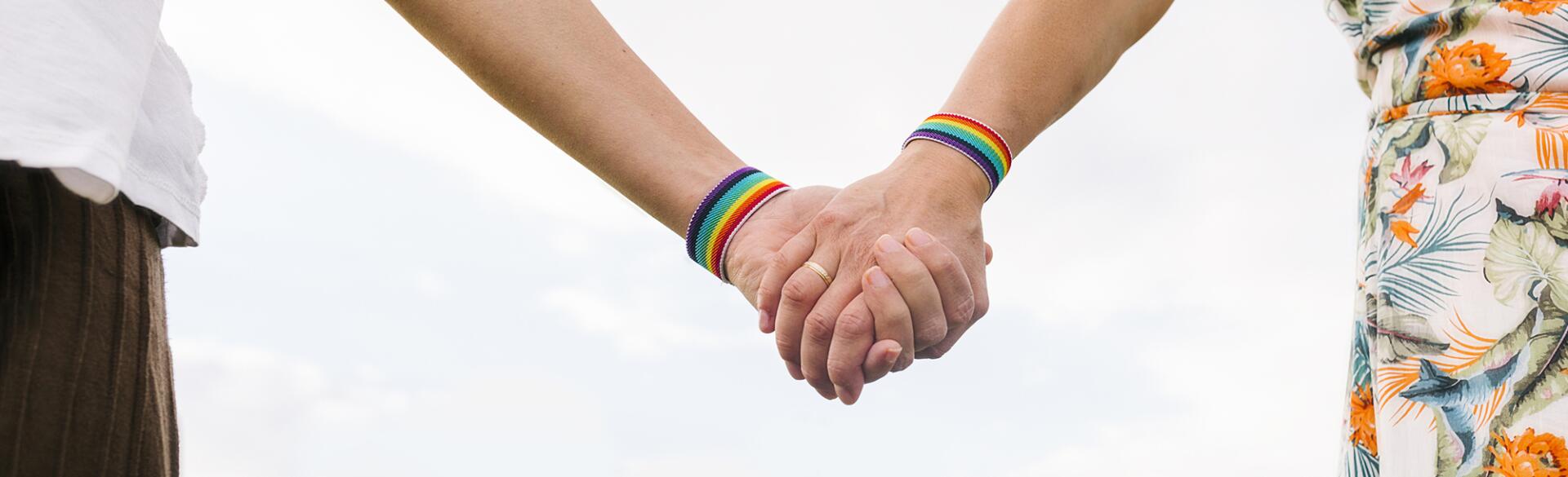 Two women holding hands each wearing a rainbow bracelet and one with a wedding band on