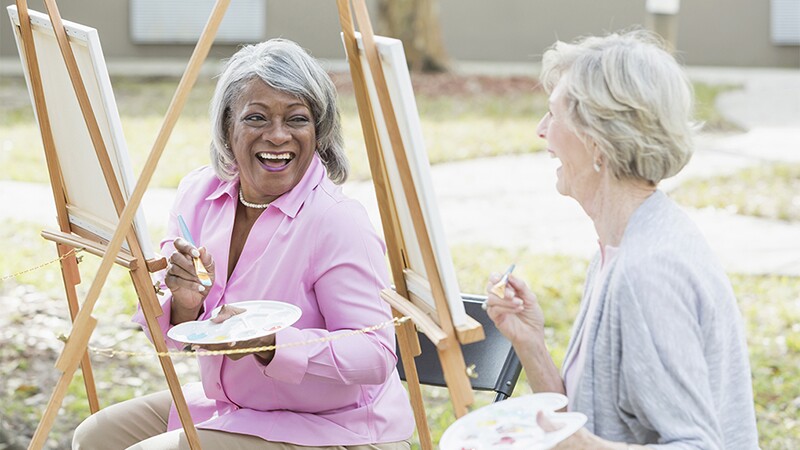 Two women smiling and laughing as they paint outdoors 