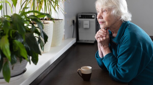 An attractive gray-haired 85-year-old elderly woman drinks espresso in the kitchen and looks out the window.