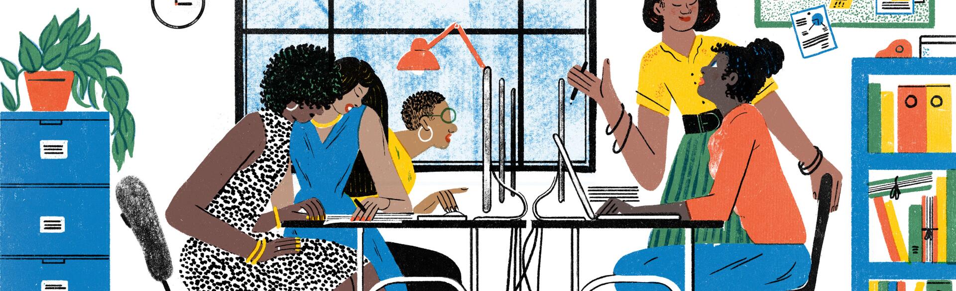 A group of women in an office space sit around a communal desk, talking and working on computers.