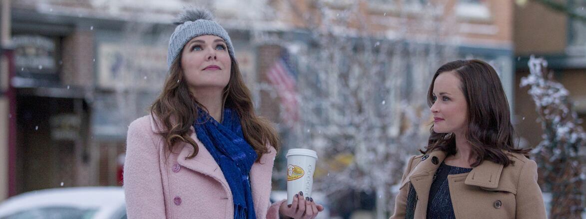 main characters from Gilmore Girls, Lorelai and Rory, outside in the snow holding coffee by a newsstand