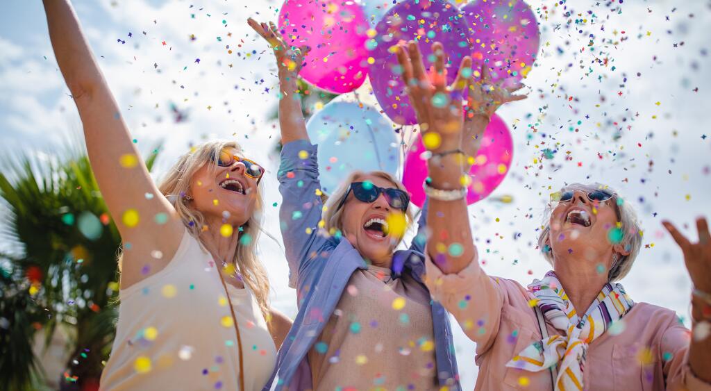 Three women celebrating with colorful confetti and balloons outdoors 
