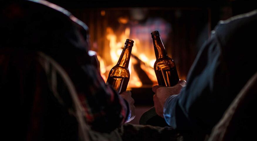 Two men rest with bottles of beer in hand by the fireplace.