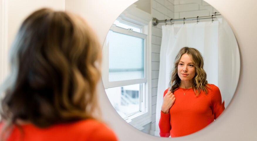 Woman in red shirt looking at reflection in bathroom mirror