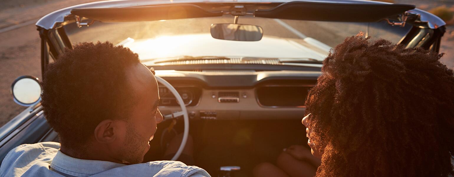 husband and wife in convertible driving into the sunset talking to each other