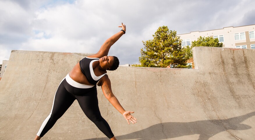 A woman stands on a ramp posing for an Instagram photo, stretching to her left side.