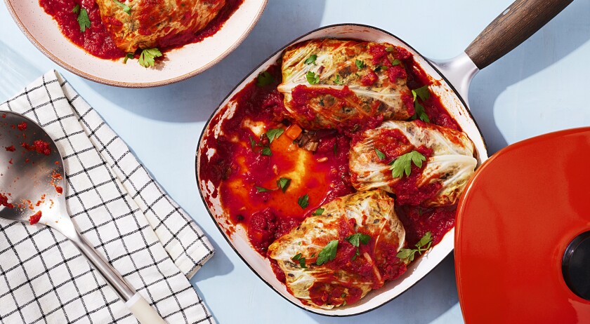 On a seamless blue surface sits a square pan with a wooden handle in the middle right of the frame. To the bottom right of the pan the bright red lid rests on the pan. Inside of the pan are three plant based stuffed cabbage rolls covered in red sauce and parsley. A forth cabbage roll covered in sauce and parsley sits half out of the frame in the top left corner on a pink speckled plate. In the bottom left corner a black and white checkered napkin or dish towel is folded and holding a used silver and white serving spoon with bits of sauce on it.