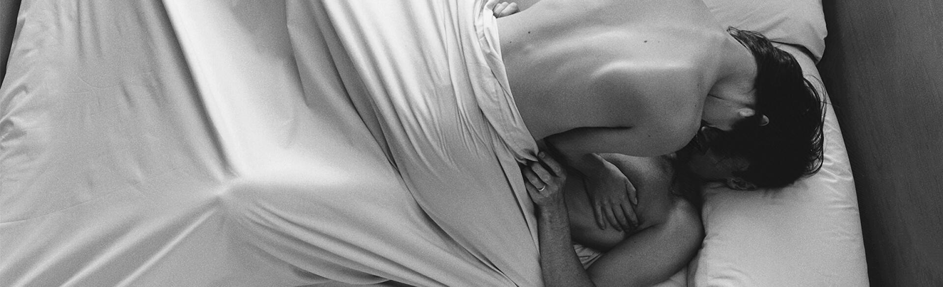 A naked woman lays on top of a man, making love in bed under a white sheet.