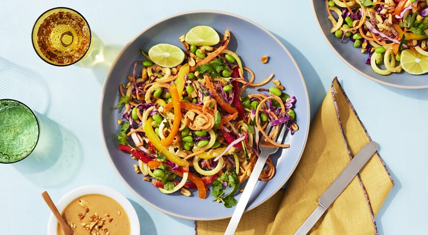 A blue dish filled with plant based zucchini noodle pad thai with carrots, peppers, beans and limes sits in the centre of the photo on a blue surface. A yellow folded napkin sits to the right of the dish along with a peanut sauce to the left of the dish. A green cup and a yellow cup with sparkling water are framed to the side and upper left of the dish. A second blue plate with pad thai is peaking into the frame in the top right corner.