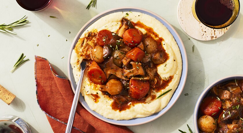 A plant based mushroom bourguignon dish with mashed potatoes, carrots and rosemary sits in the center frame inside of a blue dish with a silver fork. Top right of the dish is a clear yellow cup with red wine sitting on a pastel peach stone coaster. A blue bowl with potatoes and carrots covered in rosemary is coming into frame in the bottom right corner. A red napkin with blue checked edges is tossed to the left of the main dish. A bottle of red wine with the cork on the tabletop is coming into frame in the bottom left corner. A second glass of red wine sits to the top left of the main dish peaking into frame and rosemary sprigs are strewn about the surface.