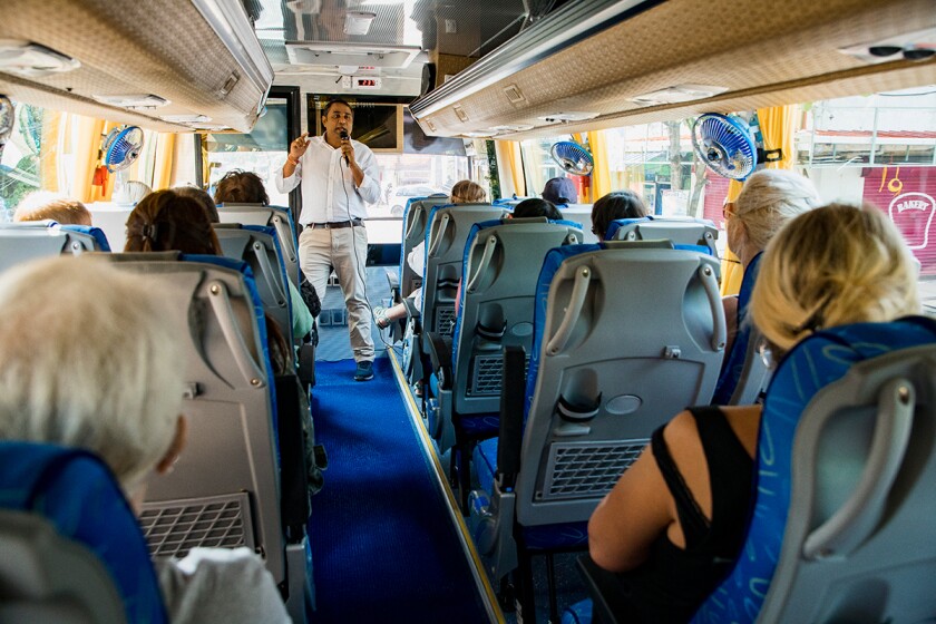 A rear-view shot of a large multi-ethnic group of tourists on a coach bus, listening to the tour guide who is speaking into a microphone