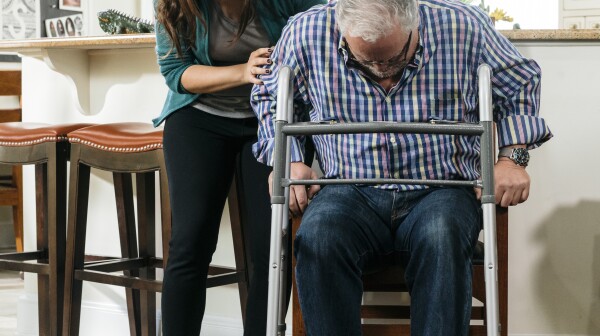 Young Hispanic woman helping her Hispanic father transfer from a seated to standing position using a walker as support.