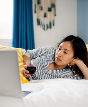 woman watching tv on her computer while laying on her bed with a glass of red wine