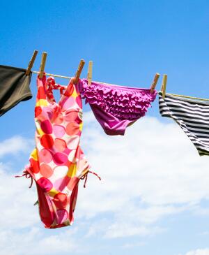 A close line with a blue sky and white clouds behind it. Bathing suits from left to right, moss green bottoms, a pink, orange, yellow and white polka dot one piece, purple ruffle bikini bottoms, black and white stripped bikini bottoms, a black bikini top, another black and white stripped bikini bottom, and a purple ruffle bikini top.