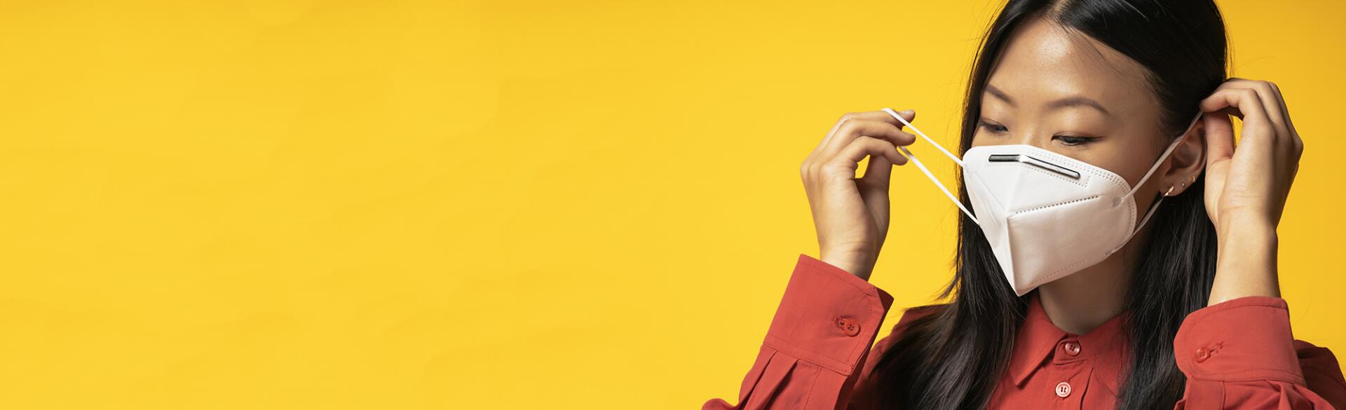 Asian Woman putting on a face mask standing in front of a bright yellow background