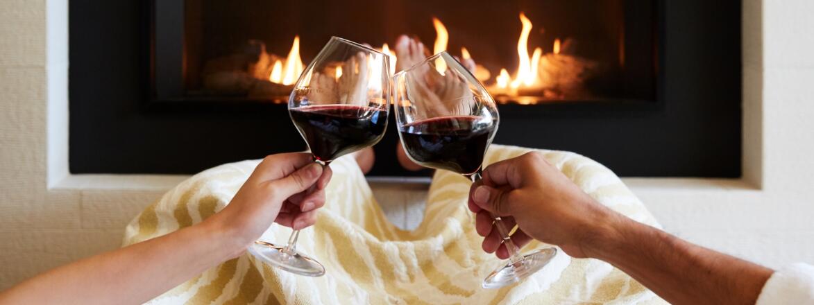 Couple with two glasses of red wine in front of a fireplace.