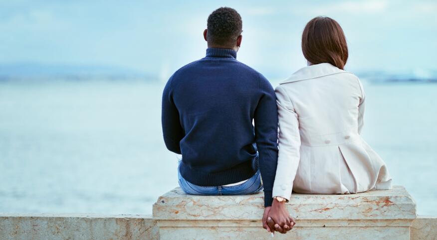 Man and woman sitting on a cement bench looking out over the ocean, holding hands