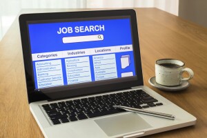 Laptop shows user interface of online job search on display screen and a cup of coffee on wooden desk, natural light coming through the window.