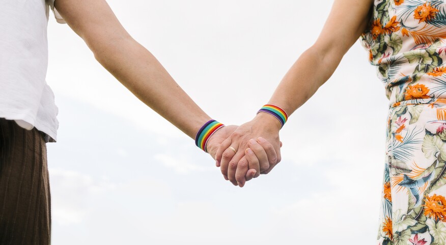 Two women holding hands each wearing a rainbow bracelet and one with a wedding band on