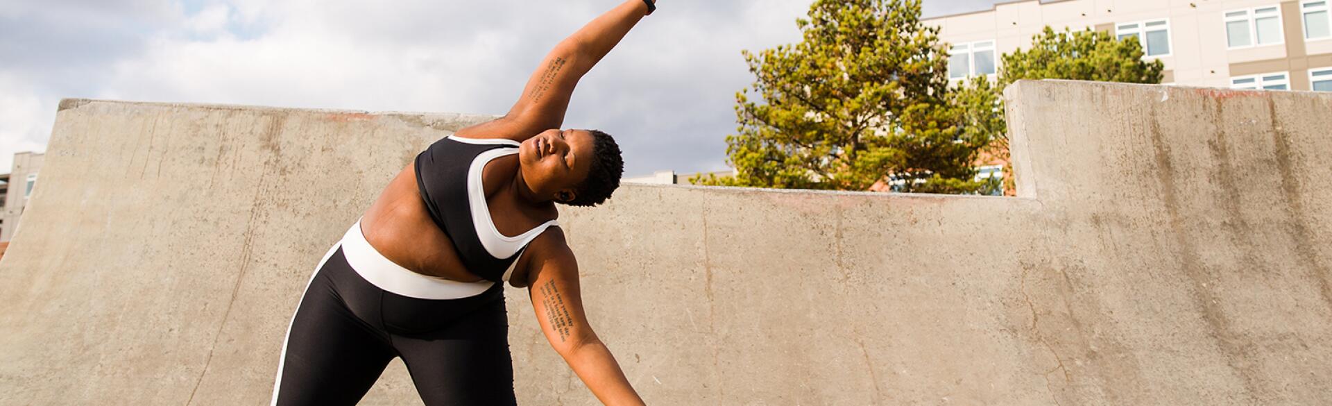 A woman stands on a ramp posing for an Instagram photo, stretching to her left side.