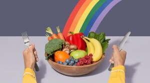 Woman with a rainbow bowl of fruits and veggies between two hands will flatware. 