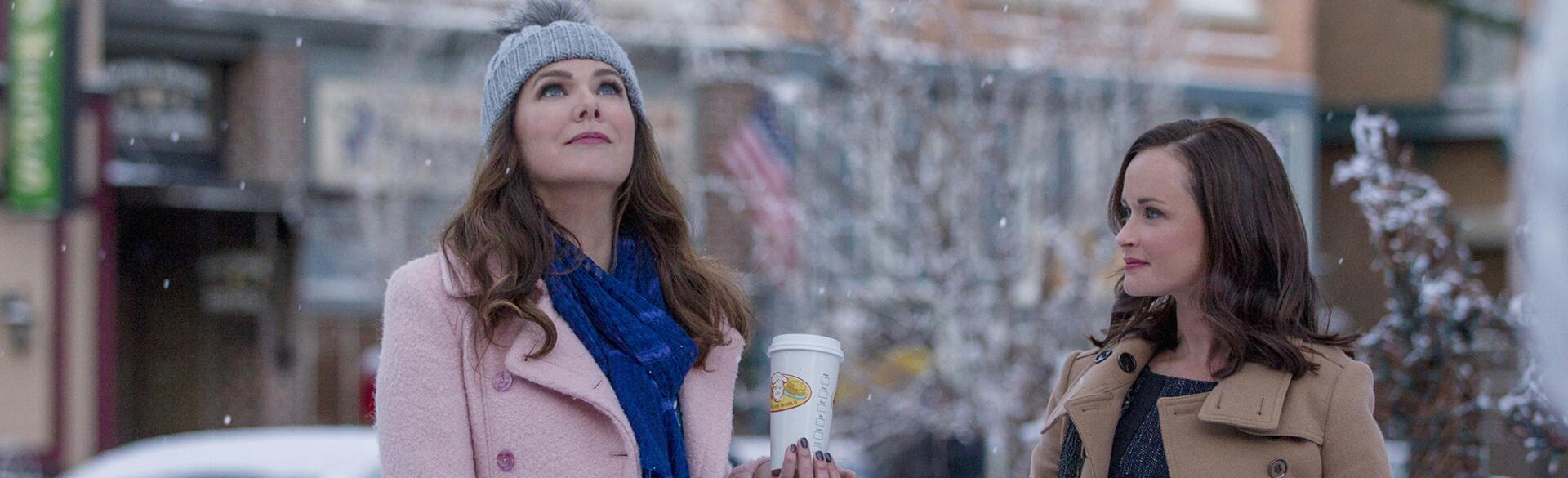 main characters from Gilmore Girls, Lorelai and Rory, outside in the snow holding coffee by a newsstand