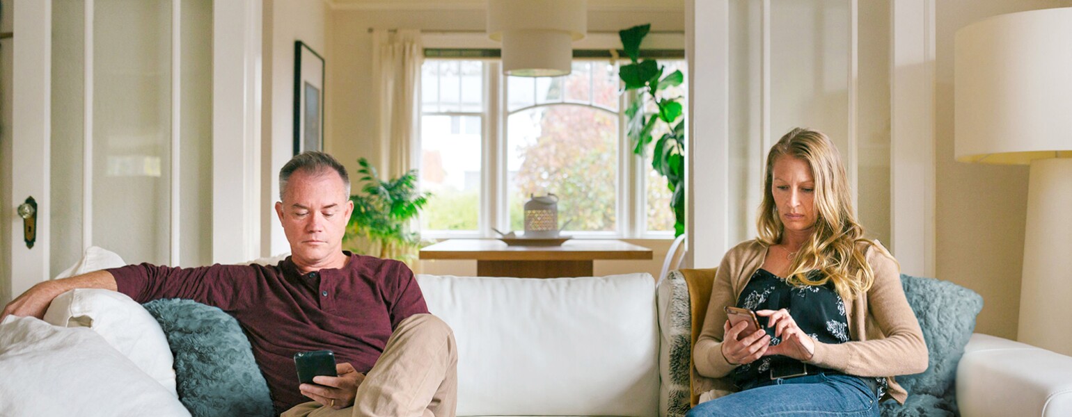 An image of a husband and wife sitting on opposite ends of the couch, distracted from each other by their cellphones.
