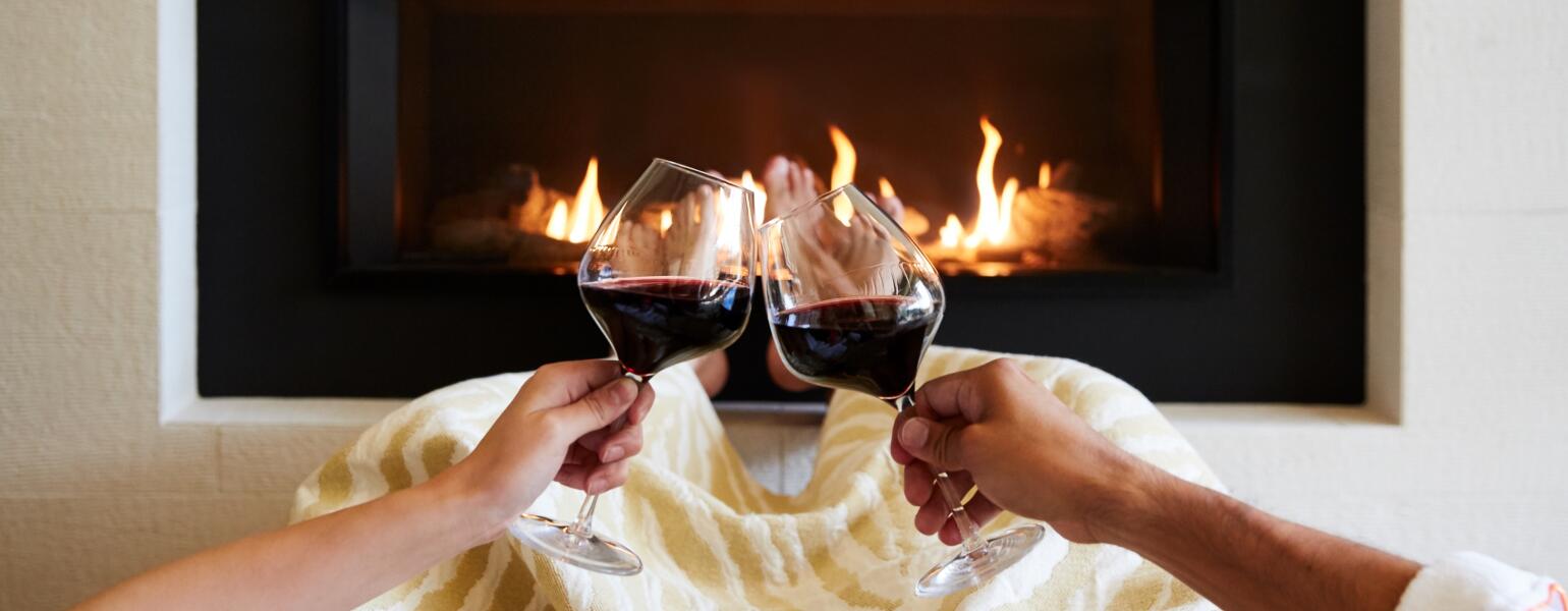 Couple with two glasses of red wine in front of a fireplace.