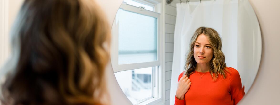 Woman in red shirt looking at reflection in bathroom mirror
