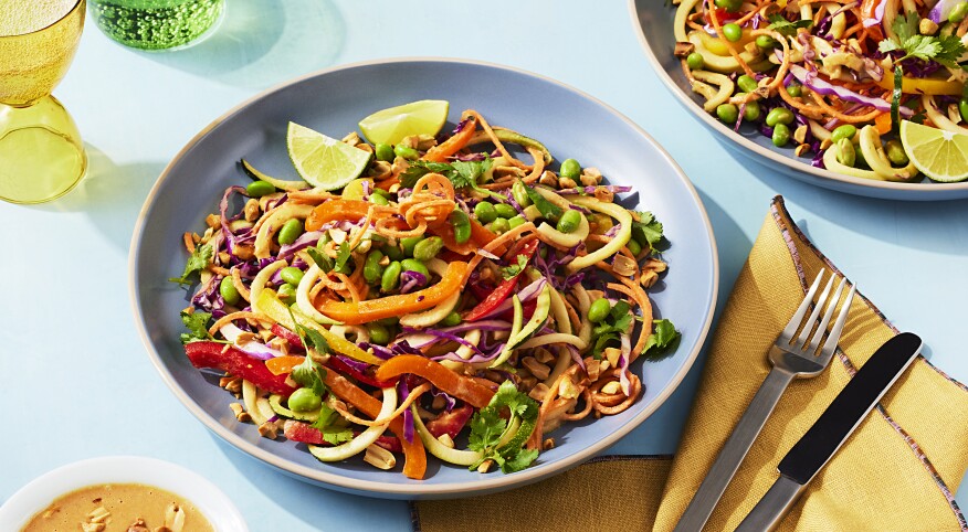 A blue dish filled with plant based zucchini noodle pad thai with carrots, peppers, beans and limes sits in the centre of the photo on a blue surface. A yellow folded napkin sits to the right of the dish along with a peanut sauce to the left of the dish. A green cup and a yellow cup with sparkling water are framed to the side and upper left of the dish. A second blue plate with pad thai is peaking into the frame in the top right corner.