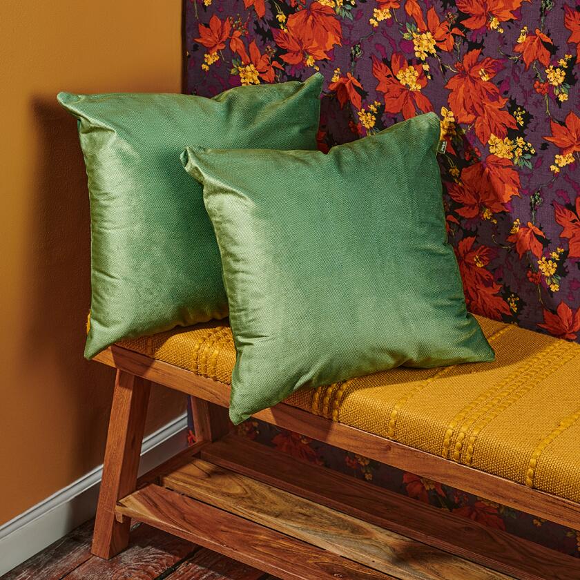 Two square green pillows propped on a bench in front of floral wallpaper