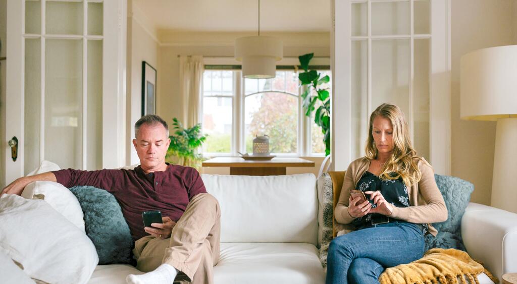 An image of a husband and wife sitting on opposite ends of the couch, distracted from each other by their cellphones.