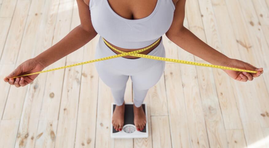 Above view of African American lady measuring her waist with tape, standing on scales indoors, closeup