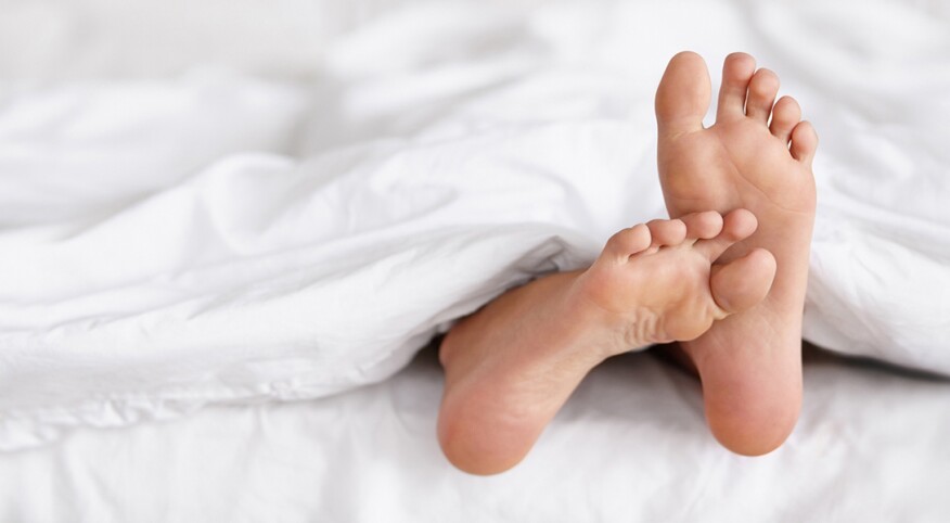 An image depicting two feet sticking out from underneath a white blanket in bed.