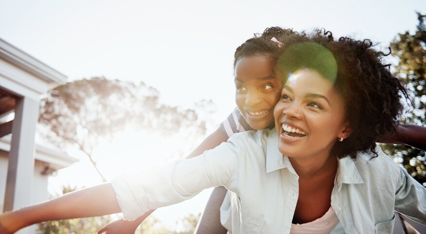 A young girl rides on the back of her aunty outside on a bright summer day.