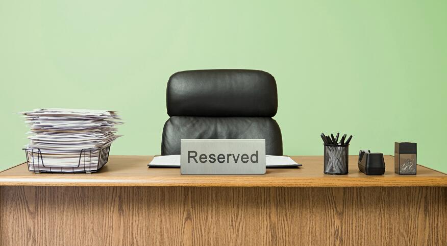 Office desk with overflowing inbox, empty desk chair with a reserved sign in front, pale green wall 