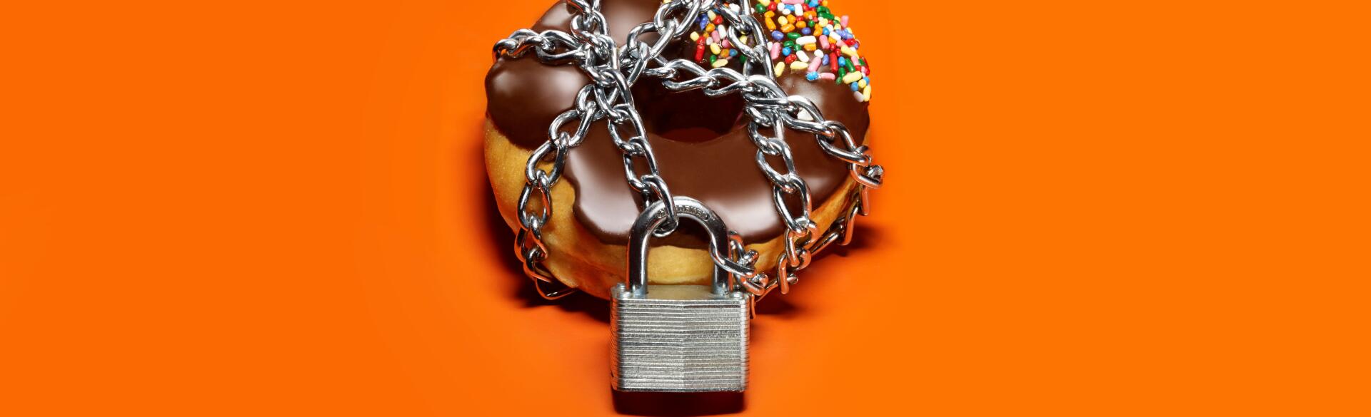 A chocolate frosted donut on an orange background with a chain and lock around it.