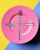 pink plate, silver fork, silver knife, plus sign on plate, yellow blue background, photograph