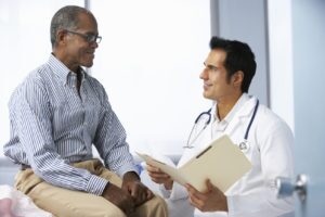 Doctor In Surgery With Male Patient Reading Notes Smiling To Each Other.