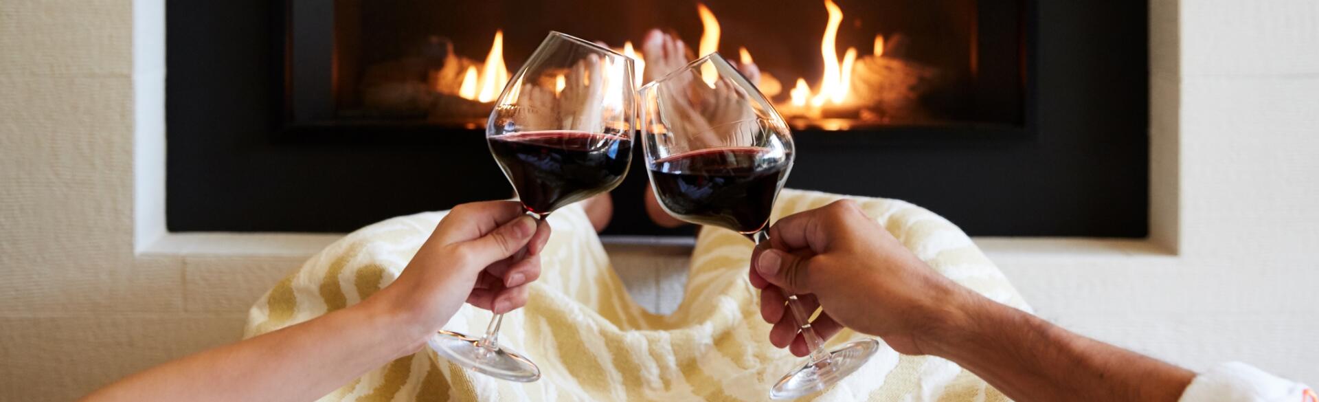 Couple with two glasses of red wine in front of a fireplace.